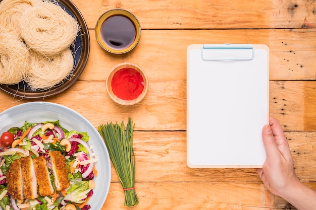 Free Photo close-up of a person's hand holding clipboard near the traditional thai food on wooden table