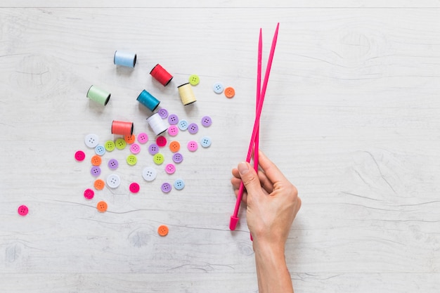 Close-up of a person's hand holding knitted needle with spool and buttons on textured backdrop