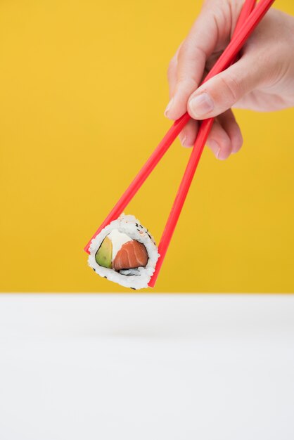 Close-up of a person's hand holding sushi roll with red chopsticks against yellow background