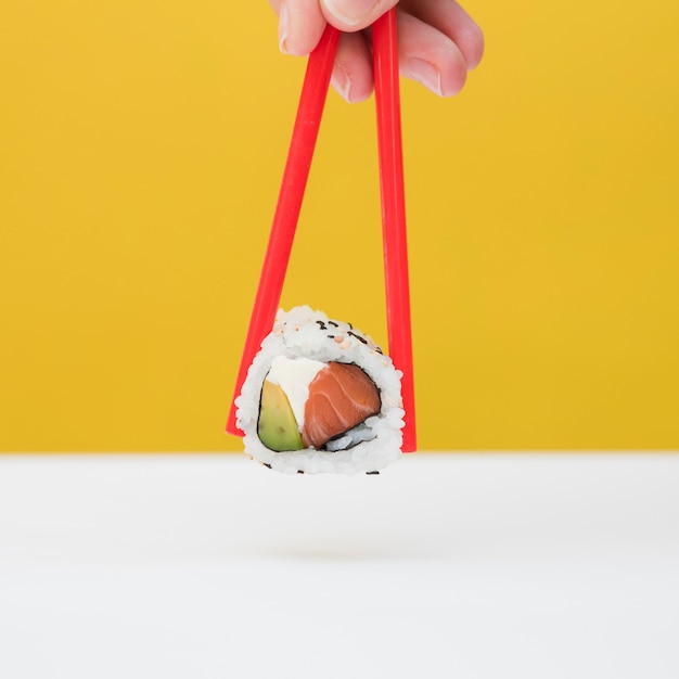 Close-up of a person's hand holding sushi with red chopsticks against yellow backdrop