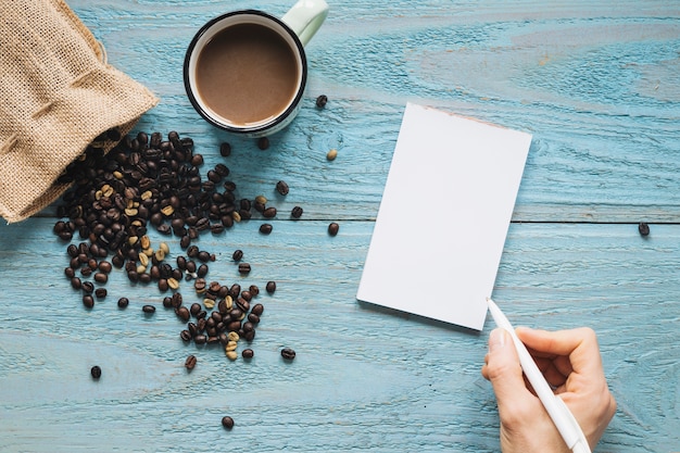 Free Photo close-up of a person's hand writing on blank paper with cup of a coffee and coffee beans