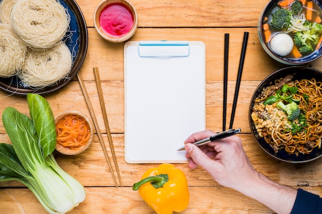 Free photo close-up of a person's hand writing on clipboard with traditional thai food on wooden table