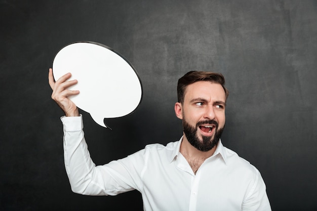 Free photo close up photo of excited man holding blank speech bubble shouting and looking aside over dark gray wall copy space