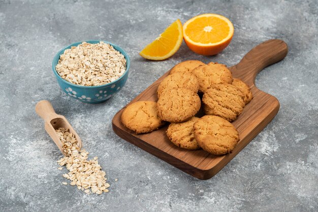 Close up photo of homemade cookies on wooden board and oatmeal with oranges over grey table.