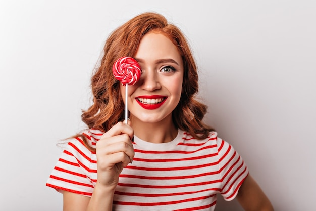 Free photo close-up photo of jocund ginger woman with red candy. joyful european girl with curly hair eating lollipop.