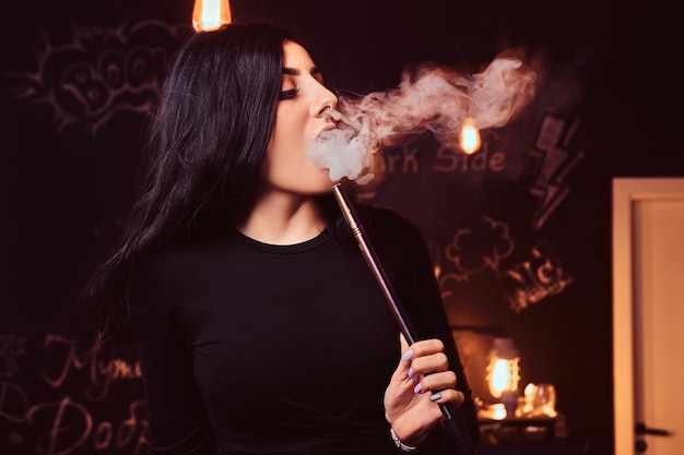 Free Photo close-up photo of a seductive brunette girl in black top smokes a hookah in a nightclub or hookah bar.