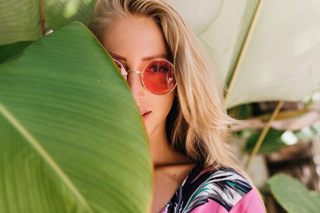 Close-up photo of sensual blonde woman in pink sunglasses hiding behind green leaf.