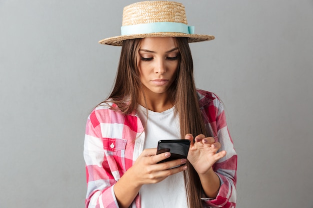 Close-up photo of serious young woman in straw hat, looking at phone,