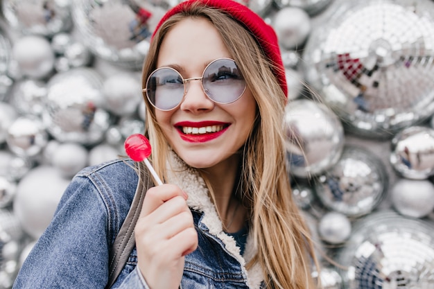 Free photo close-up photo of smiling girl in denim jacket posing with red lollipop. portrait of amazing white female model standing near disco balls with candy.