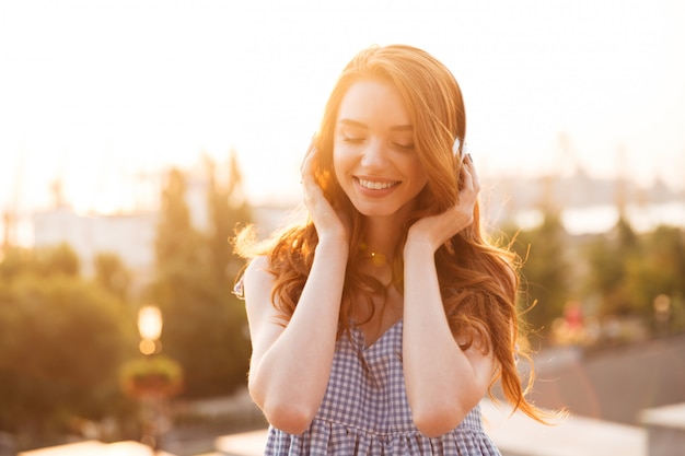 Close up picture of Attractive ginger woman in dress listening music on the sunset and looking down