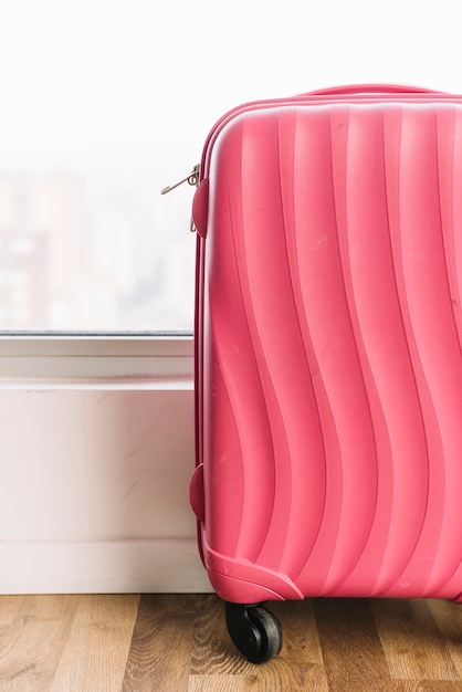 Free photo close-up of pink travel bag with wheels on wooden floor near window