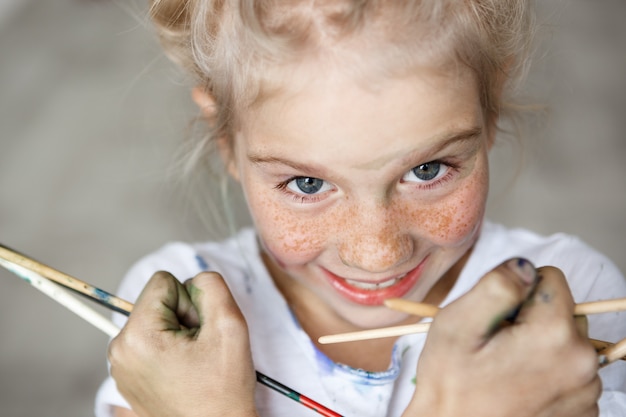 Free photo close up portrait of adorable blonde little female child in white t-shirt holding brushes, having fun, enjoying drawing with happy expression