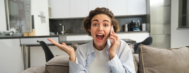 Free Photo close up portrait of amazed brunette woman jumps on sofa from excitement answers phone call hear