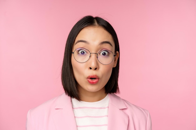 Close up portrait of asian businesswoman in glasses looking surprised at camera amazed reaction standing in suit over pink background