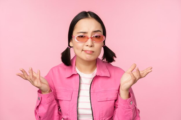 Close up portrait of asian girl looking confused shrugging puzzled and looking at camera wearing sunglasses standing over pink background