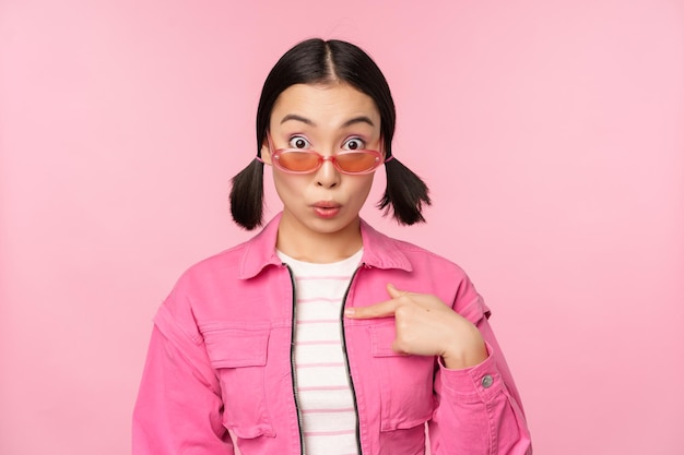 Close up portrait of asian girl looks surprised points at herself with disbelief being chosen stands over pink background Copy space