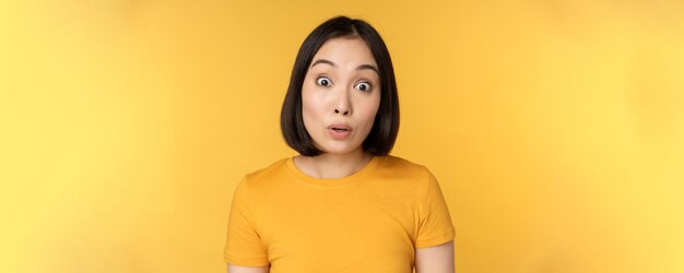 Close up portrait of asian girl showing surprised reaction raising eyebrows amazed reacting to big news standing over yellow background