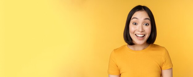 Close up portrait of asian girl showing surprised reaction raising eyebrows amazed reacting to big news standing over yellow background