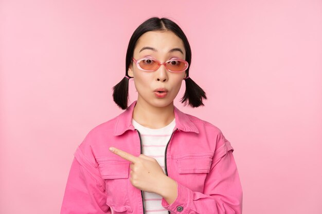 Close up portrait of asian woman gasping looking surprised pointing finger at banner advertisement standing over pink background