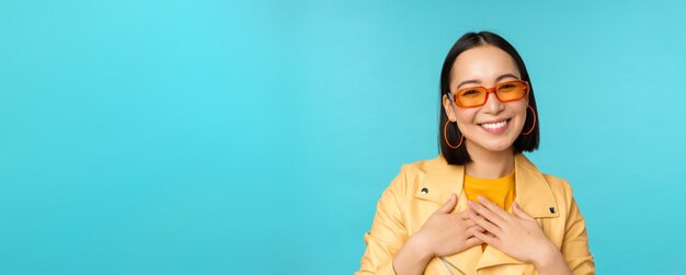 Close up portrait of asian young woman in sunglasses smiling and looking romantic standing happy over blue background Copy space