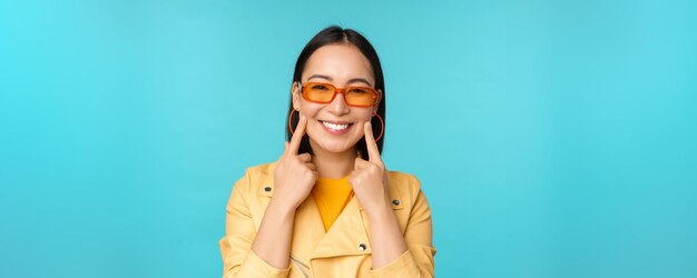 Close up portrait of asian young woman in sunglasses smiling and looking romantic standing happy over blue background