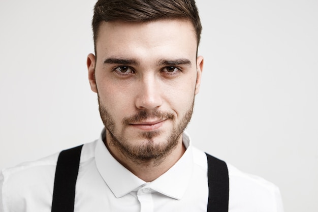 Close up portrait of attractive young brunette man with trendy hairstyle and trimmed beard posing at white studio wearing formal shirt and suspenders, feeling confident about upcoming job interview