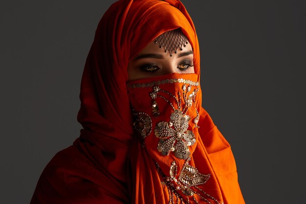 Close-up portrait of an attractive young female with beautiful smoky eyes and jewelry on the forehead, wearing the terracotta hijab decorated with sequins. She is posing and looking down on a dark bac