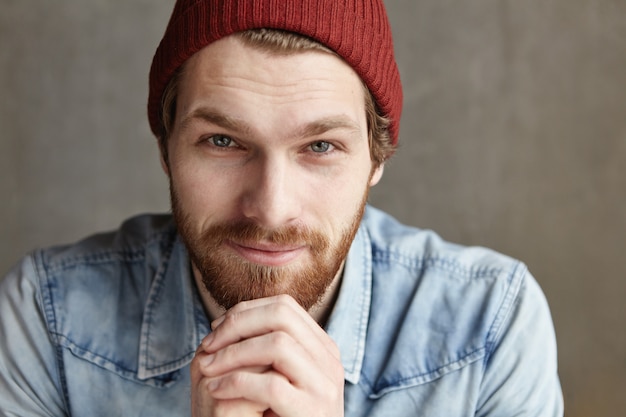 Free photo close up portrait of attractive young male with thick beard and charming blue eyes wearing stylish clothing, looking with flirty smile, keeping hands on his chin. people and lifestyle