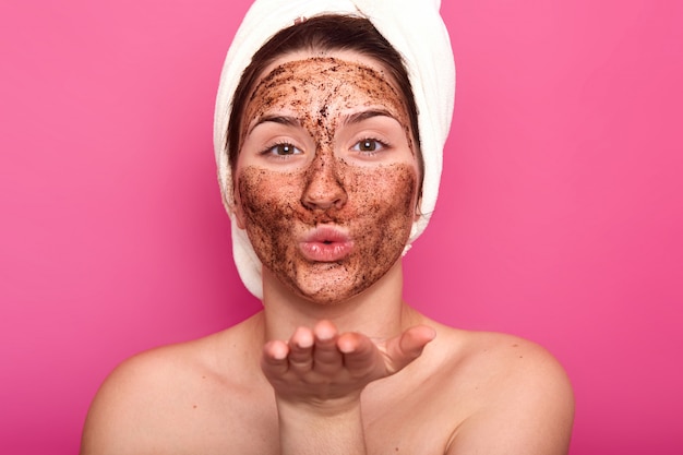 Close up portrait of beatiful European female blowing kiss, putting chocolate face mask, being naked, coning her hair with white towel, looks peaceful and relaxed. Beauty and care concept.