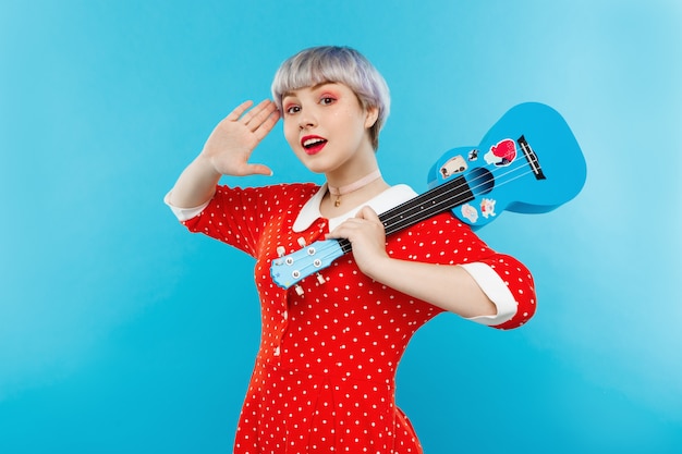 Free photo close up portrait of beautiful dollish girl with short light violet hair wearing red dress holding ukulele over blue wall