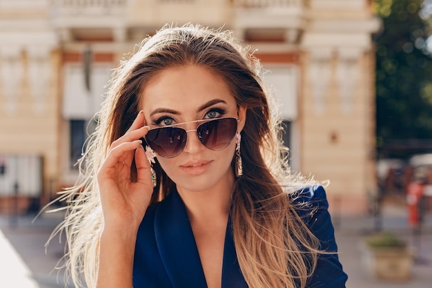 Close-up portrait of beautiful woman dressed in stylish blue jacket walking in autumn sunny street wearing elegant sunglasses