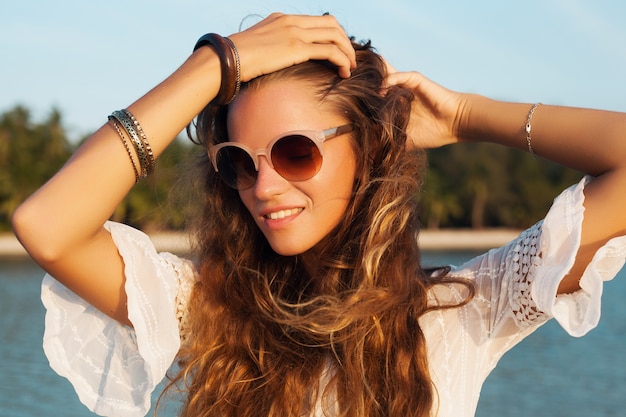 Close up portrait of beautiful woman in white dress on tropical beach on sunset wearing stylish sunglasses.