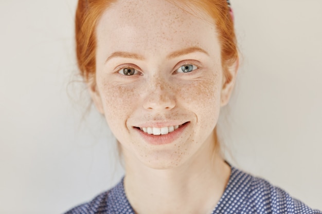 Free Photo close up portrait of beautiful young redhead model with different colored eyes and healthy clean skin with freckles smiling joyfully, showing her white teeth, posing indoors. heterochromia in human