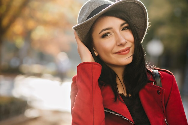 Close-up portrait of black-haired woman with nude makeup posing with pleasure on blur forest background. Smiling lady in hat and red coat expressing true emotions.