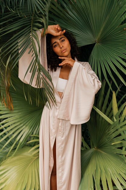 Close up portrait of blissful woman with wavy hairs posig in tropical garden Wearing elegant silk robe Spa and skincare concept