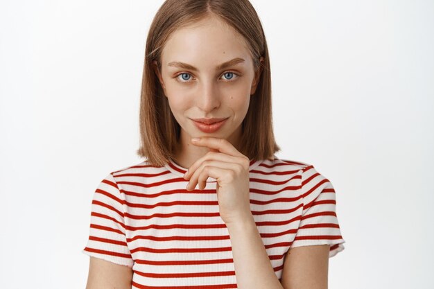 Close up portrait of blond woman clean natural face without makeup, looking thoughtful and determined, smiling and staring as if having an idea, great plan, standing against white wall