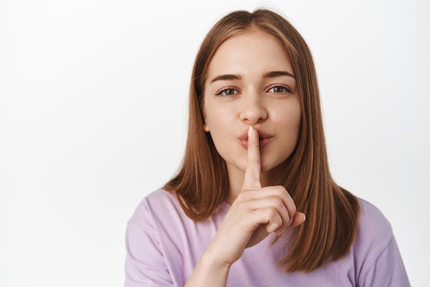 Free Photo close up portrait of blond woman shushing, smiling and asking to keep quiet, has secret, shhh gesture, press finger to lips, has surprise, standing against white wall