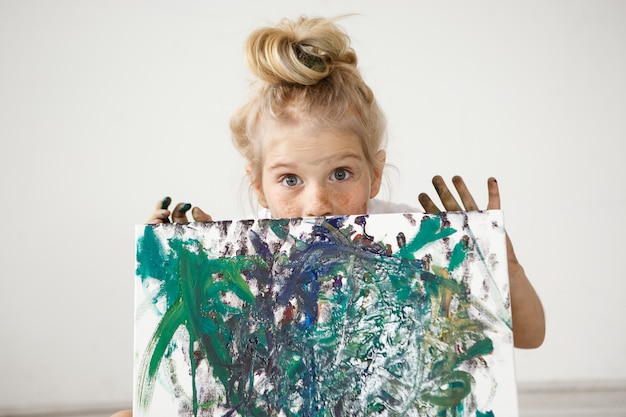 Free photo close-up portrait of blonde european little girl with hair bun and big blue eyes demonstrating her picture.