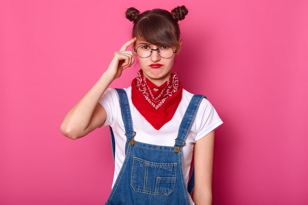 Free photo close up portrait of brunette student, wears round eyewear and red bandana on neck, curves lips, keeps fore finger on temple