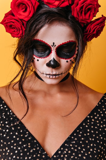 Free photo close-up portrait of brunette with crown of roses humbly bowing her head. woman with skull makeup lowered her slight.