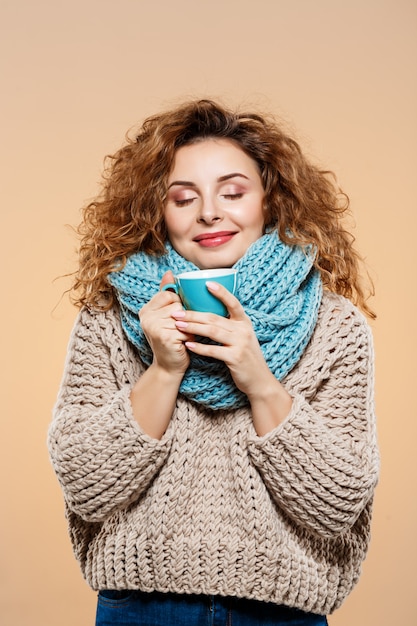 Free photo close up portrait of cheerful smiling beautiful brunette curly girl in knitted sweater and grey neckwarmer holding cup over beige wall