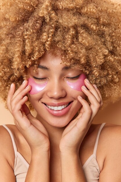 Close up portrait of cheerful woman with curly hair smiles happily applies hydrogel patches under eyes for reducing wrinkles keeps eyes closed enjoys beauty treatments. Skin care and rejuvenation