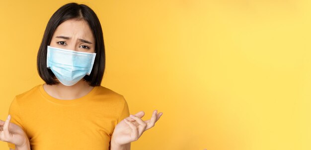 Close up portrait of confused asian woman in medical face mask shrugging shoulders and looking puzzl