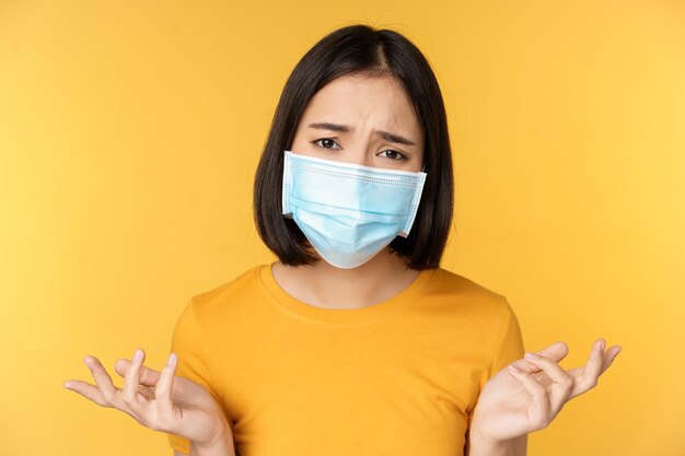 Close up portrait of confused asian woman in medical face mask shrugging shoulders and looking puzzled standing against yellow background