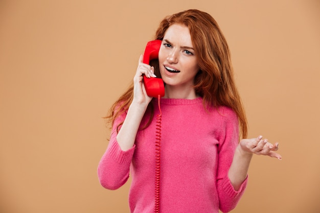 Close up portrait of a confused pretty redhead girl talking by classic red phone