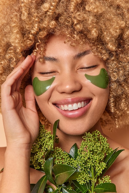 Close up portrait of curly haired woman smiles broadly shows white teeth applies green patches for reducing puffiness undergoes beauty procedures keeps eyes closed. Women wellness skin care.