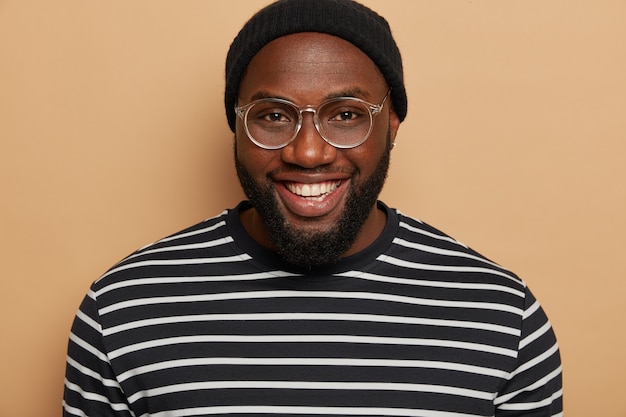 Close up portrait of dark skinned adult man with thick bristle, smiles toothy, wears big optical glasses, striped jumper, glad to meet friend