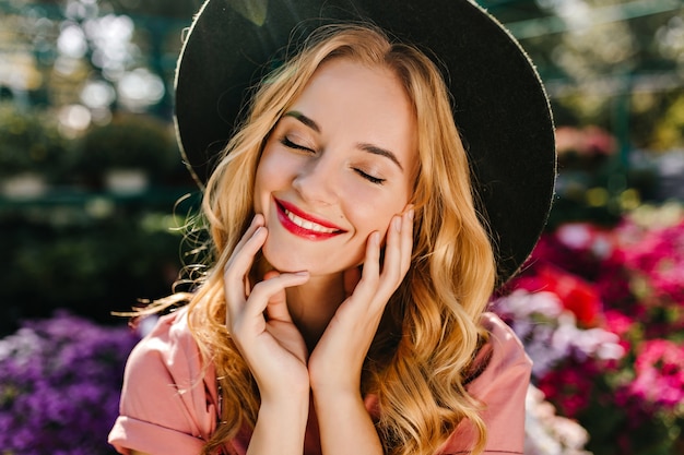 Close-up portrait of ecstatic blinde woman with red lips wears black hat. Debinair white woman posing with eyes closed near flowers.