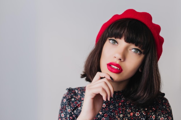 Free photo close-up portrait of elegant brunette girl with short hair wearing red beret and touching her chin. beautiful young dark-haired woman in vintage outfit posing meditatively isolated on light background