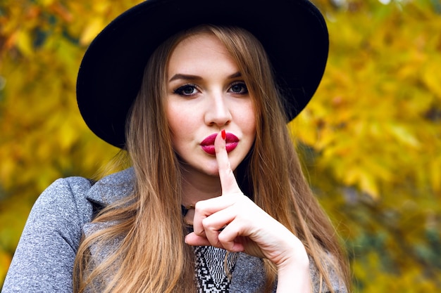 Close up portrait of elegant pretty blonde woman posing at autumn cold day in the city park, wearing elegant black hat, long hairs, bright make up